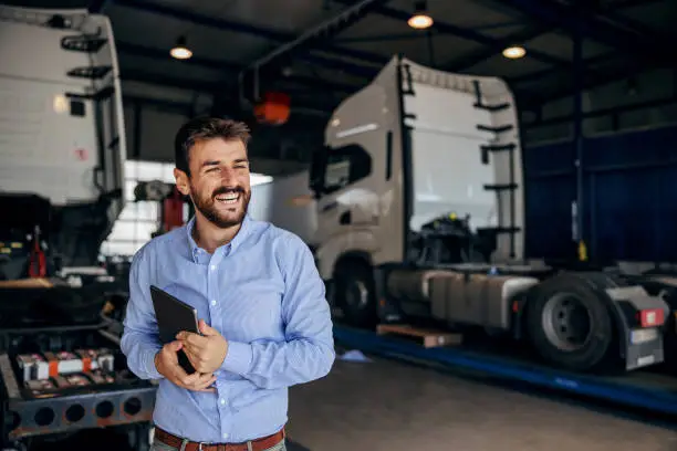 Smiling chief standing in auto park and holding tablet. In background are trucks. Firm for import and export.