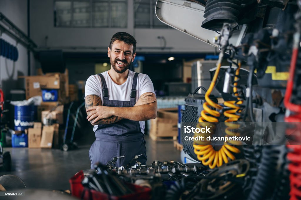 Smiling happy bearded tattooed worker in overalls standing next to truck with arms crossed. Mechanic Stock Photo