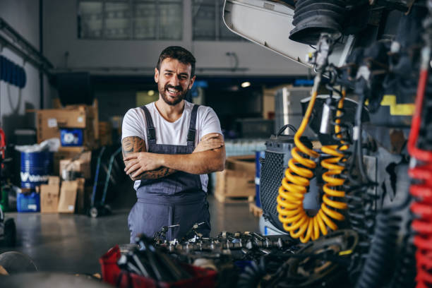 smiling happy bearded tattooed worker in overalls standing next to truck with arms crossed. - machine part imagens e fotografias de stock