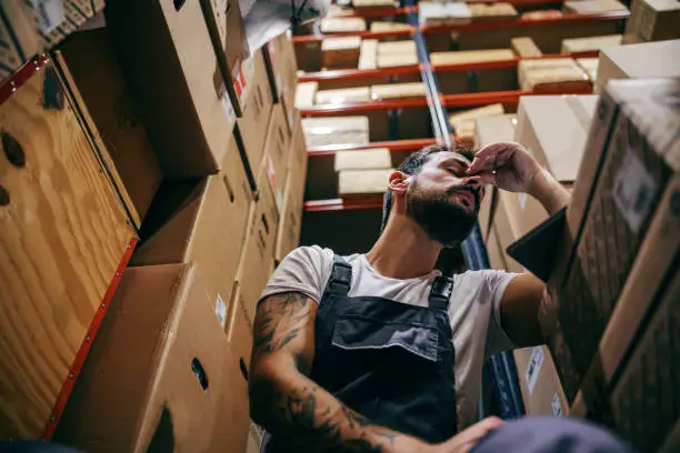 Photo of Tattooed bearded worker in overalls having hard day on the work. Storage of import and export firm interior.