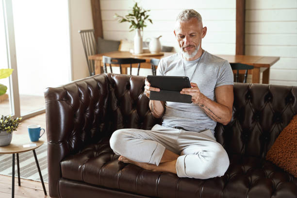 istock Middle aged caucasian man in casual clothes using digital tablet while sitting in lotus pose on sofa in the living room of his modern apartment 1285074317