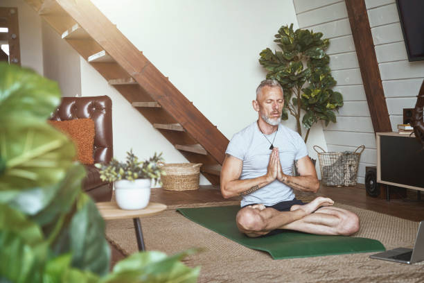 Healthy and calm middle aged man sitting in lotus pose in the living room and meditating, watching online yoga class on laptop Healthy and calm middle aged man sitting in lotus pose in the living room and meditating, watching online yoga class on laptop. Sport and active lifestyle during self isolation man yoga tattoo stock pictures, royalty-free photos & images