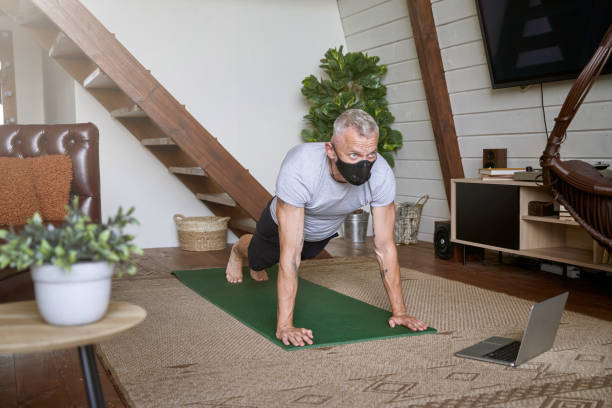 istock Sporty middle aged man wearing face protective mask standing in plank pose while exercising at home during self isolation, watching online workout on laptop 1285074462