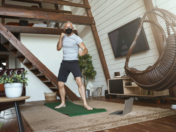 istock Covid 19 outbreak. Active middle aged man in face protective mask standing in boxing stance in the living room at home, watching online workout on laptop 1285074551