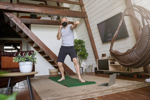 Sport during quarantine. Active middle aged man in face protective mask standing in boxing stance in the living room at home, watching online workout on laptop
