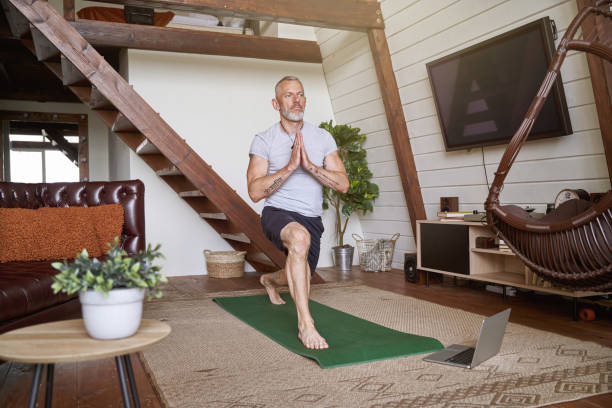 istock Healthy and calm middle aged man practicing yoga at home while watching online video tutorial on laptop, exercising in the living room 1285074562