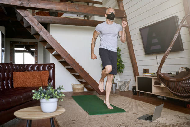 istock Sporty middle aged man wearing face protective mask exercising in self isolation while watching online workout on laptop in the living room of his modern apartment 1285074581