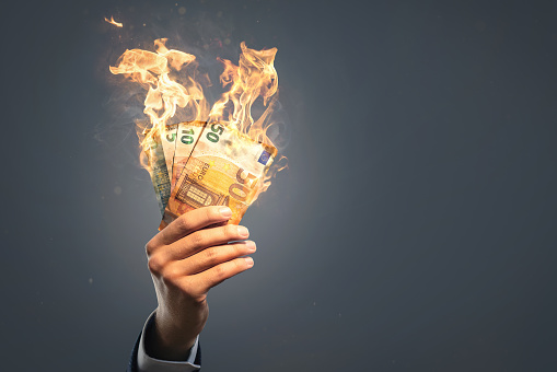 A hand of a businessman is holding several burning Euro banknotes. Copy space and neutral background.