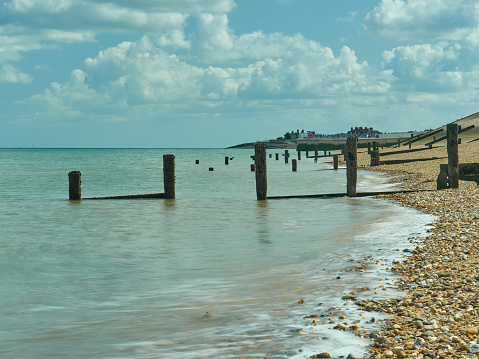 Landscape / seascape image taken at Sheerness-on-Sea. Image shows a wide-angle view along the waterline. To the right quarter, the image shows the pebbles of the shingle beach. The left three quarters show a motion-blurred sea, softened by a long exposure. The image is punctuated by decaying sea-defences, broken wooden groynes, with an outcrop of buildings on the horizon under a blue summer sky, piled up with cotton-wool clouds.