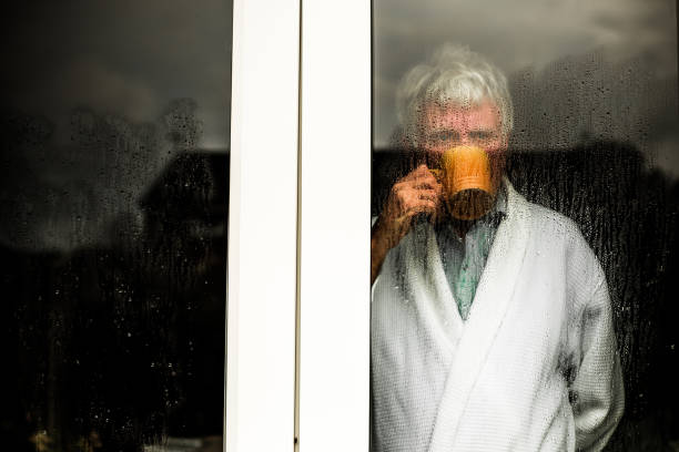 Close up desaturated color image depicting a sad, depressed-looking senior man in his 60s, and of caucasian ethnicity, looking of a rain-soaked window. The man is wearing a dressing gown and holding a cup of coffee. The image was taken during the Covid-19 pandemic during lockdown, and the image illustrates a mental health concept. Room for copy space.