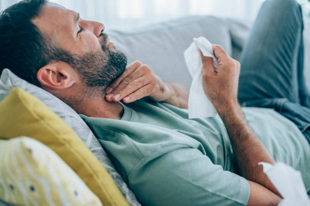 Illness young man with throat pain lying on the sofa at home. Sick man lying on the couch with high temperature and holding his throat with hand. sore throat stock pictures, royalty-free photos & images