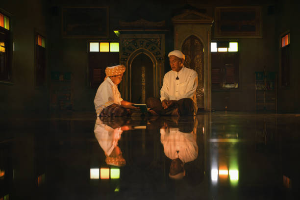 мусульманский мальчик, сидящий в темной мечети с исламским учителем, изучая книгу корана во время рамадана - изучая стоковые фото и изображения