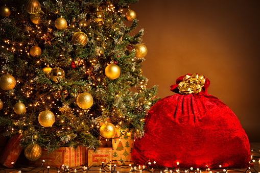 Santa Red Bag full with Gifts. Christmas tree Decorated by Golden Balls and Presents Box under. Big Xmas Sack tied by Golden Ribbon over Brown Background Interior