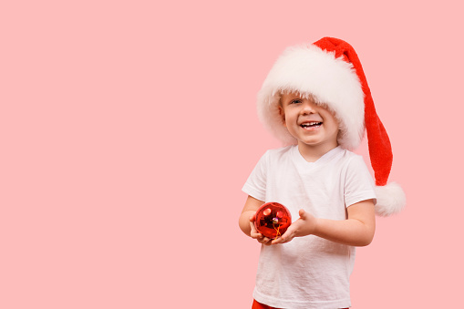 Boy child Toddler kid 3 years old in a hat of Santa Claus stands and holds a New Year's ball. Studio shot concept, postcard on a pink background close up with place for text and congratulations.