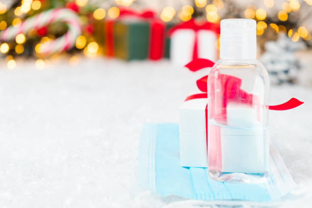 istock Gift box with a red bow from ribbon, sanitizer bottle in the protective face mask against coronavirus on the background of presents under Christmas tree. Christmas and New Year holidays during pandemic of Covid-19 1285096430