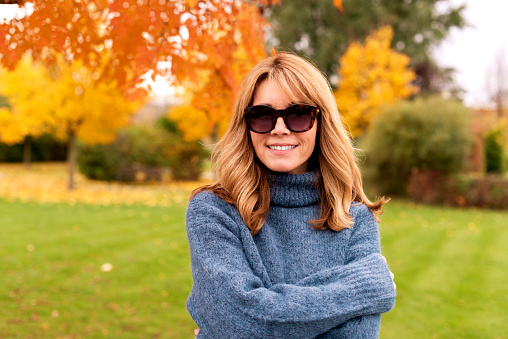 Portrait shot of smiling mature woman wearing sunglasses and turtleneck sweater while relaxing at the park in autumn.