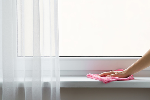 Young adult woman hand wiping white plastic window sill from dust with pink dry rag. Daily home chores. General or regular cleanup. Closeup. Empty place for text on white glass. Copy space.