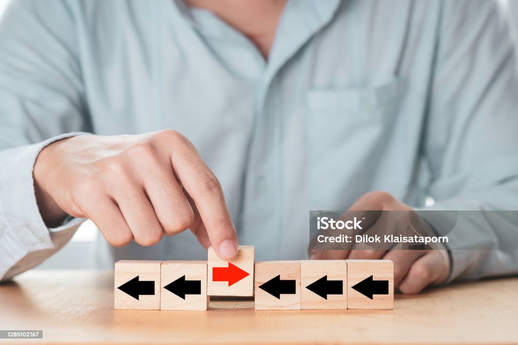 Disruption  and different thinking concept , businessman holding red arrow among black arrow which print screen on wooden cube block and it opposite direction. Arrow Symbol Stock Photo