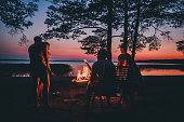 Group of young happy friends sitting by the fire at summer beach, grilling sausages and drinking beer, talking and having fun