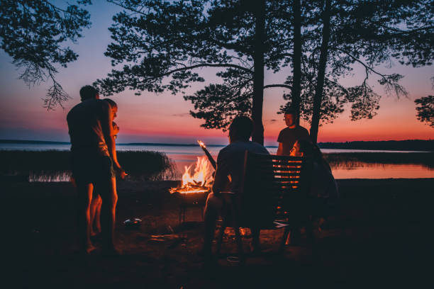 Photo of Group of young happy friends sitting by the fire at summer beach, grilling sausages and drinking beer, talking and having fun
