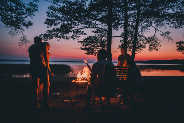 group of young happy friends sitting by the fire at summer beach, grilling sausages and drinking beer, talking and having fun - picnic summer break relaxation imagens e fotografias de stock