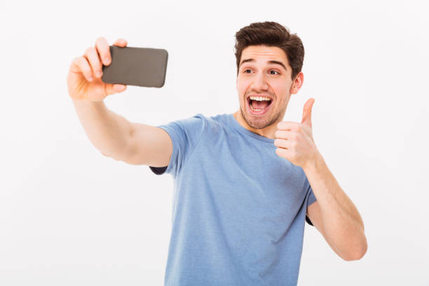 istock Image of happy guy in studio smiling and showing thumb up while photographing himself on black smartphone, isolated over white background 1285112628