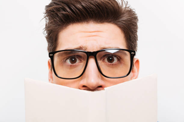 istock Close-up image of Confused nerd in eyeglasses hiding behind book 1285116957