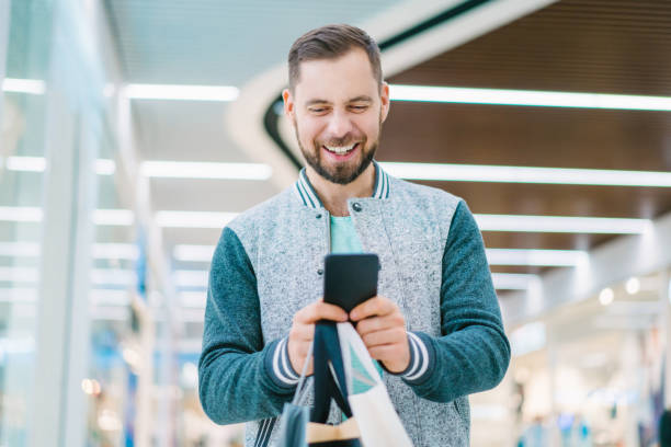 손에 쇼핑 가방의 무리와 쇼핑몰에서 걷는 동안 자신의 휴대 전화를 사용하여 캐주얼 옷을 입고 젊은 수염남자를 미소 잘생긴 미소 - mouth open retail shock surprise 뉴스 사진 이미지