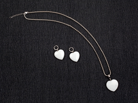 Pair of white heart shaped agate earrings and pendant with silver chain on black textured background. Close-up shot