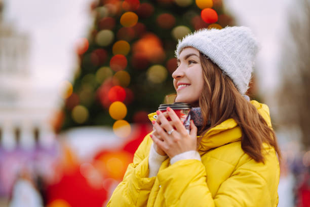 Young woman drinking hot coffee while walking near New year tree. Young woman drinking hot coffee while walking near New year tree. Happy woman wearing knitted hat and scarf, holding thermos with hot tea in frosty winter day. Festive Christmas fair, winter holidays new year urban scene horizontal people stock pictures, royalty-free photos & images