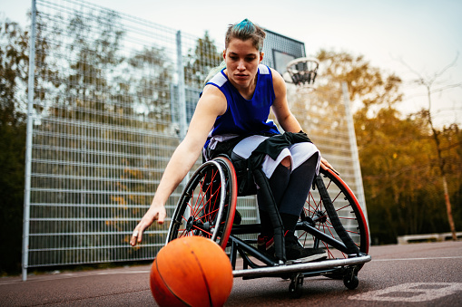 Disabled 20 y.o. woman in wheelchair, shooting basketball ball on the outdoor court on rainy day