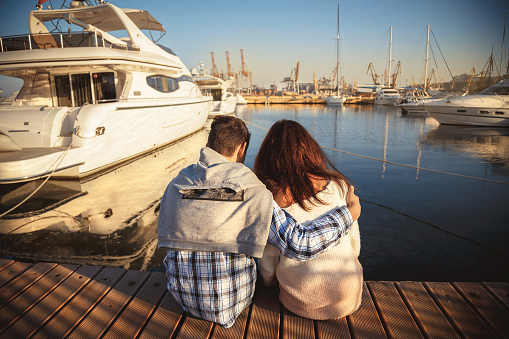 Back view of young couple sitting together on wooden pier in the port with small yachts near to the sea water