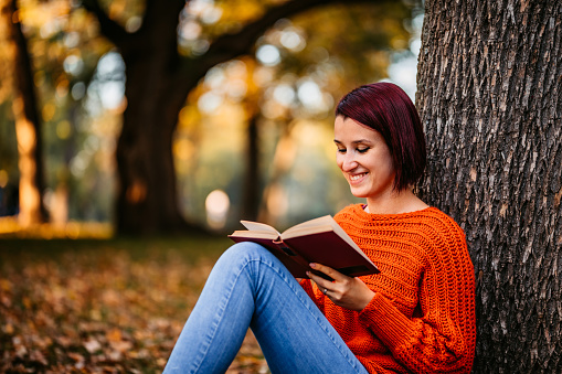 Beautiful young woman reading book under tree in public park.