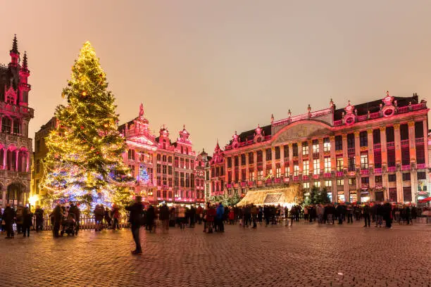 Photo of Christmas tree in a crowded square surrounded by illuminated historic buildings at night. Brussels, Belgium.