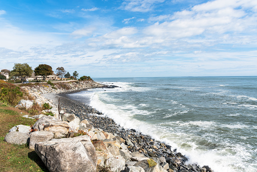 View of a small pebble beach along the coast of New Hampshire on a sunny autumn day. A clifftop coastal road lined with houses is visible in background. Portsmouth NH, USA.