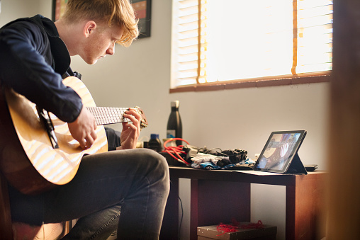 Teenage boy learning to play the guitar with an online class on a digital tablet during lockdown at home