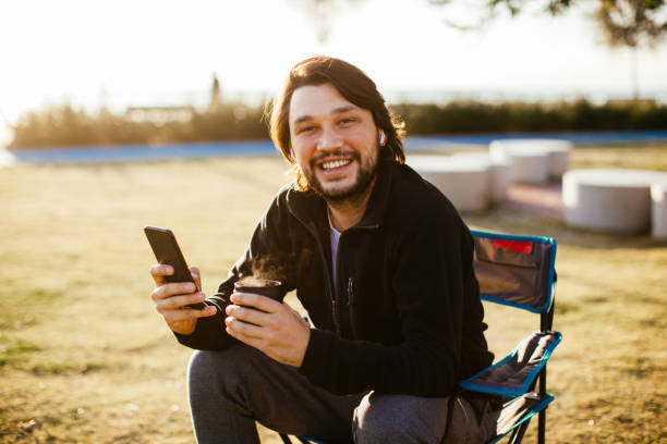 homme s’asseyant sur la chaise de camp et utilisant le téléphone portable - fresh coffee audio photos et images de collection