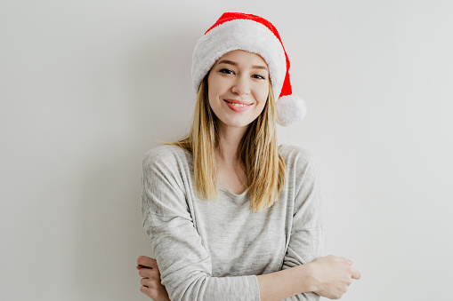 A young smiling woman stands in front of a white wall and has a Santa Claus hat on her head