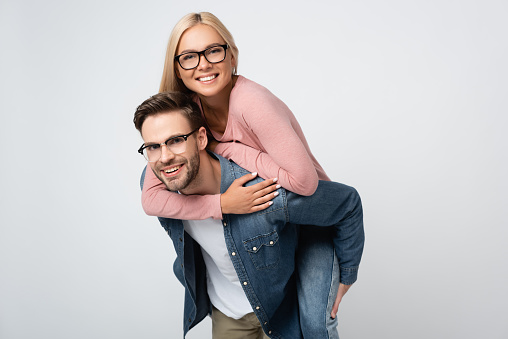 Smiling woman in eyeglasses piggybacking on boyfriend isolated on grey