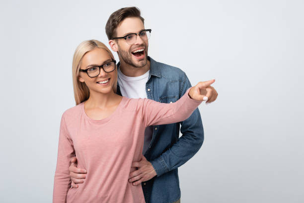 Cheerful man in eyeglasses embracing girlfriend pointing with finger isolated on grey