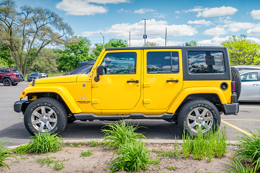 A yellow colored Jeep Wrangler Sahara Unlimited suv is parked in a parking lot in Burlington, Ontario, Canada on a sunny day. Side view.