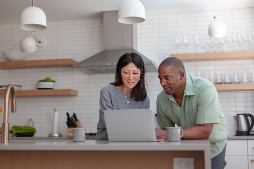 A mixed race couple use a laptop computer to pay bills online, manage budget and prepare tax documents. They are in the kitchen of their home.