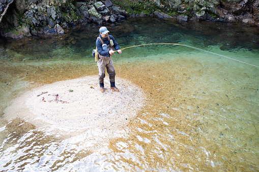A Japanese man Fly Fishing in a small mountain river with a waterfall behind him.