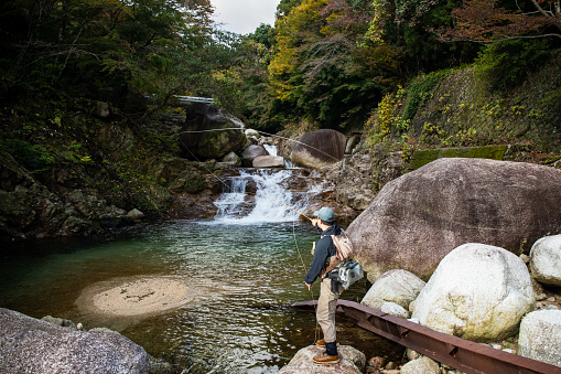 A Japanese man Fly Fishing in a small mountain river with a waterfall behind him.