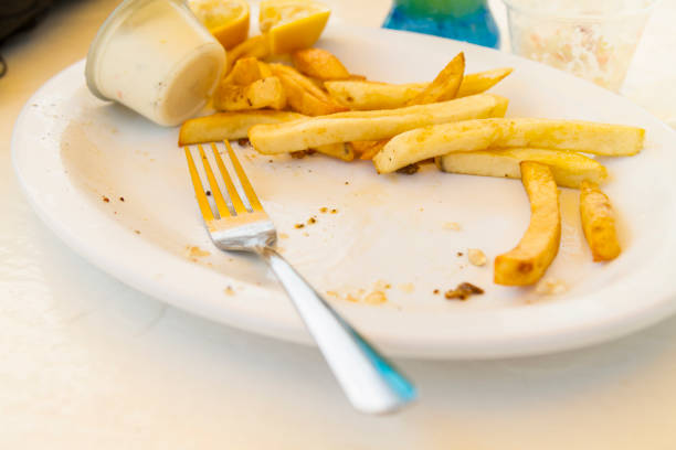 Scraps on an empty greasy plate with leftover Fish, chips and coleslaw food over a plastic table at the beach, on left a metal shinny fork.