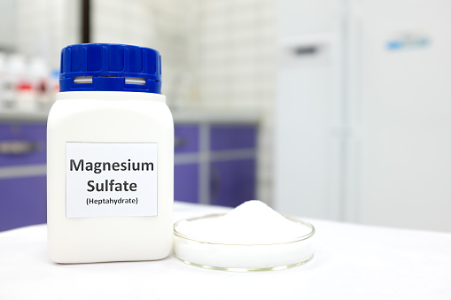 Selective focus of magnesium sulfate or epsom salt chemical compound bottle beside a petri dish with white crystalline powder in a chemistry laboratory background.