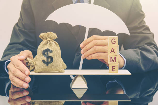 Businessman holds an umbrella to protect a US dollar money bag and cubes with a word GAME on a simple balance scale The image depicting balancing between money and a business which could be success and failure ponzi scheme stock pictures, royalty-free photos & images