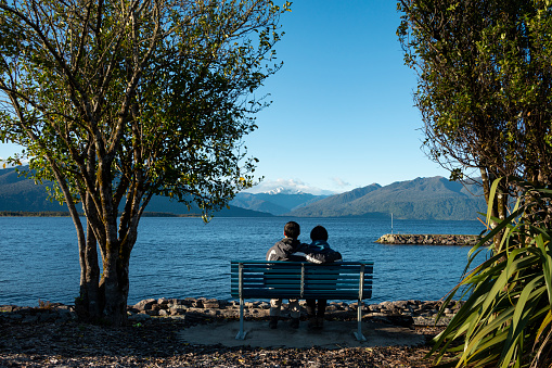 Couple sitting on the bench and enjoying the views of snow-capped mountains at Lake Brunner, South Island, New Zealand