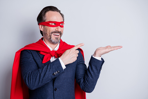 Photo of confident cheerful aged mature business guy super hero costume, direct fingers open arm hold novelty product empty space offer wear suit red face mask cloak isolated grey background