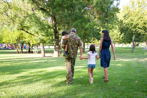 Back view of happy family walking together on meadow in park. Father wearing camouflage uniform, holding son and enjoying weekend with wife and kids. Family reunion and returning home concept